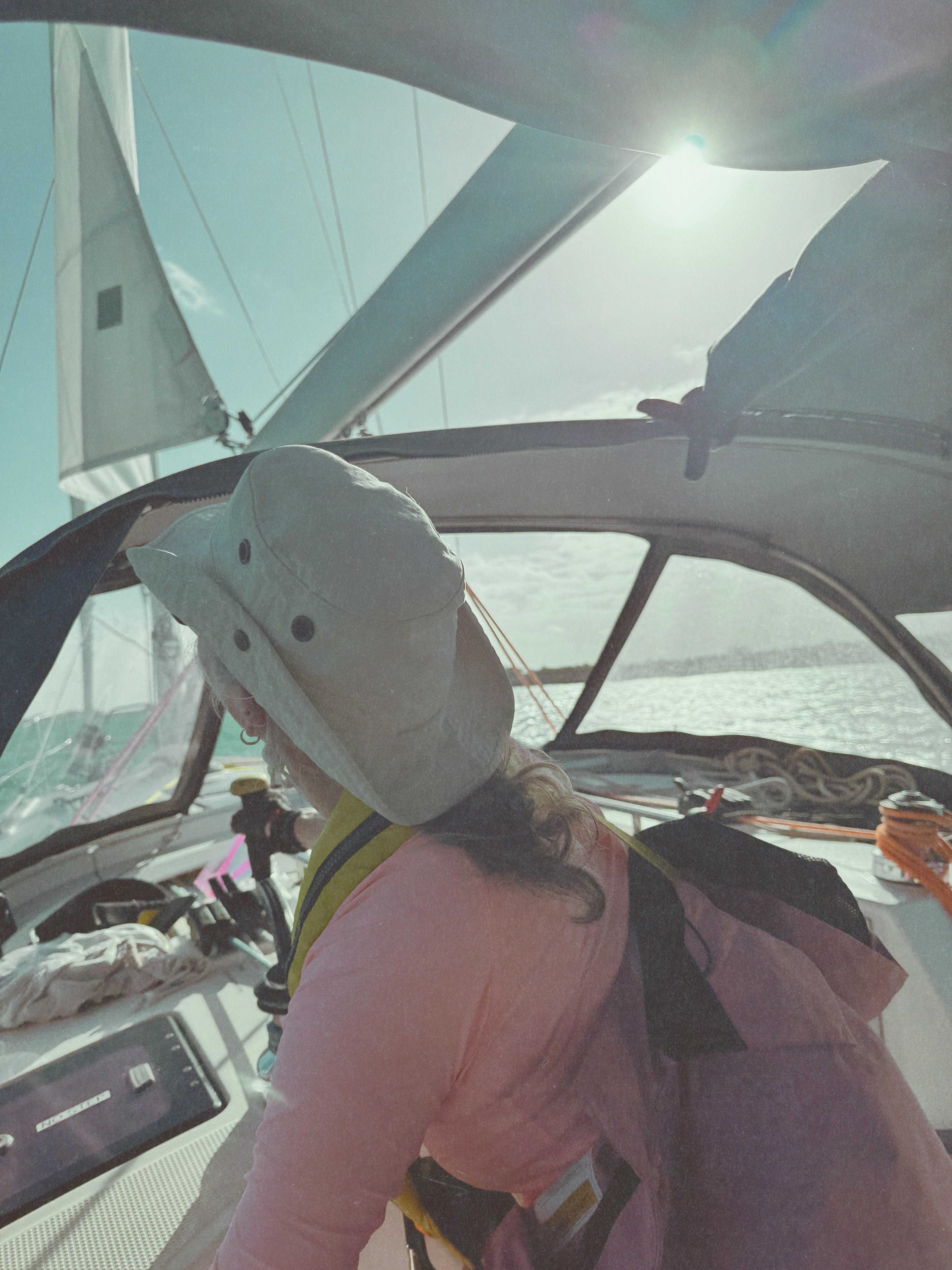 Beth at the helm of a cruising sailboat in the Exumas, backlit by the midday sun through the mainsail.