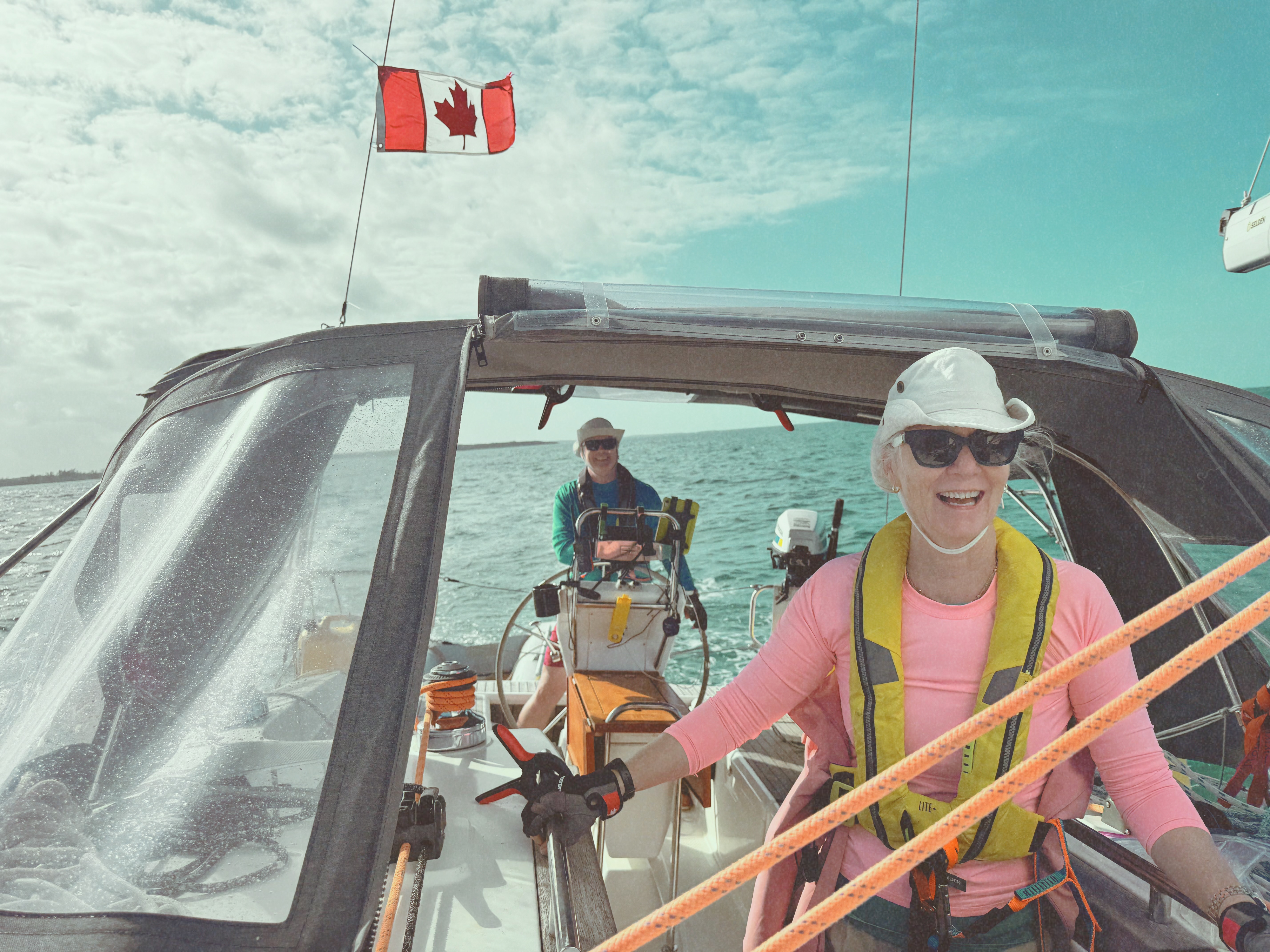 A Canadian flag flying astern of a cruising sailboat in the Exumas, with a smiling sailor in a life jacket at the foredeck.