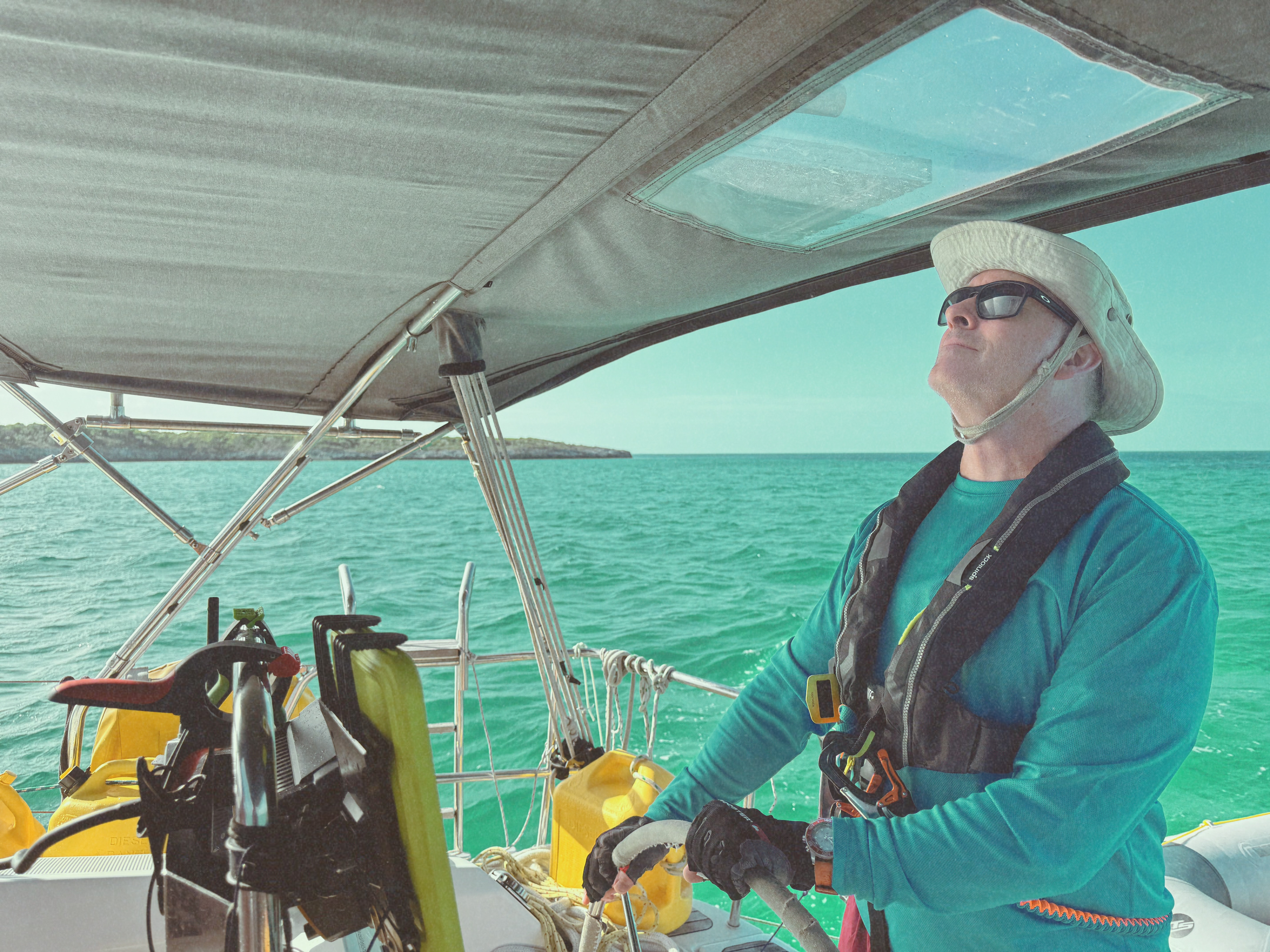 Ian at the wheel of a cruising sailboat under the bimini, looking up to read the shape of the mainsail.