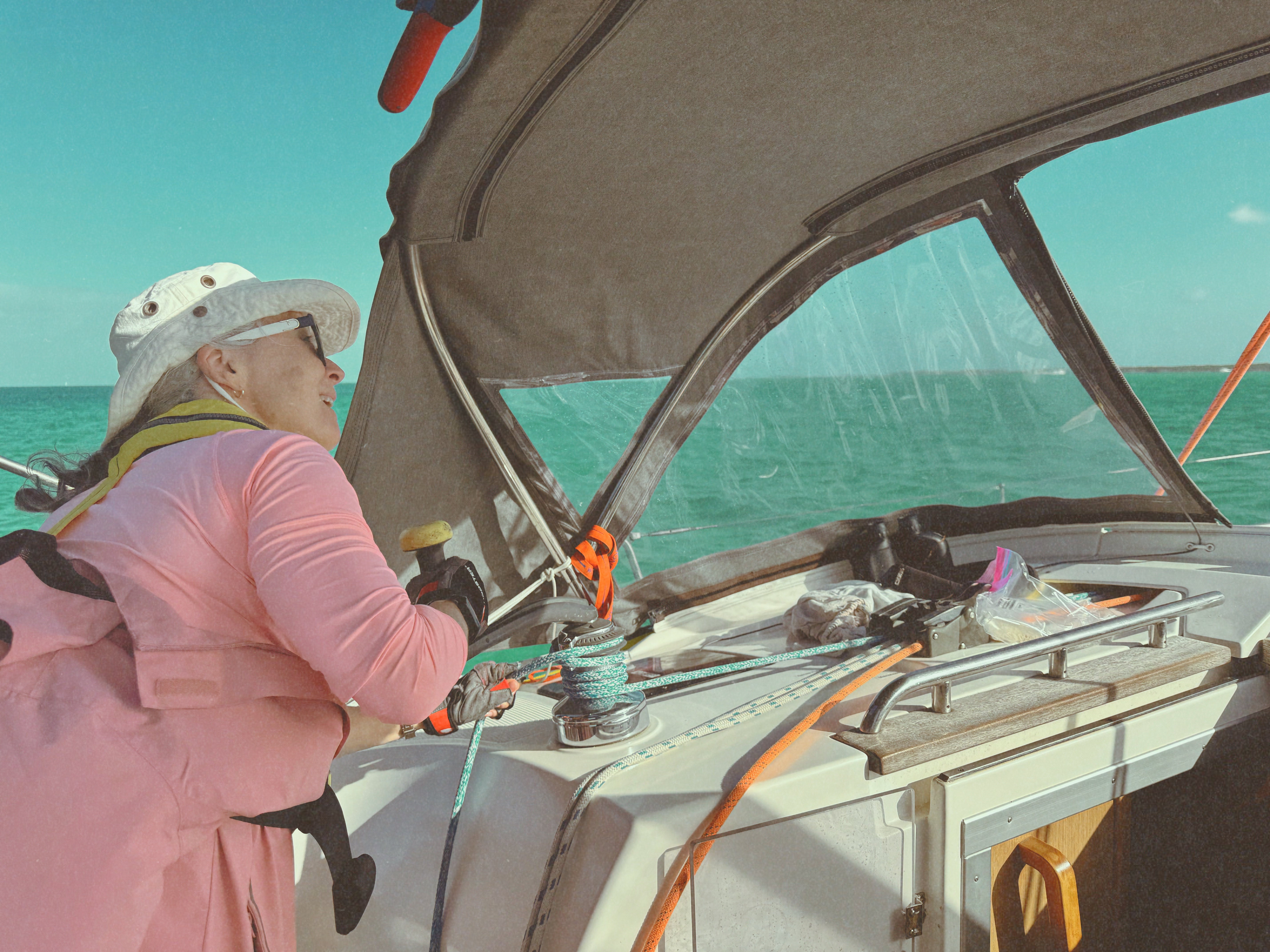 Beth in a pink jacket at the cockpit winch of a cruising sailboat, looking forward over the turquoise Bahamian bank.