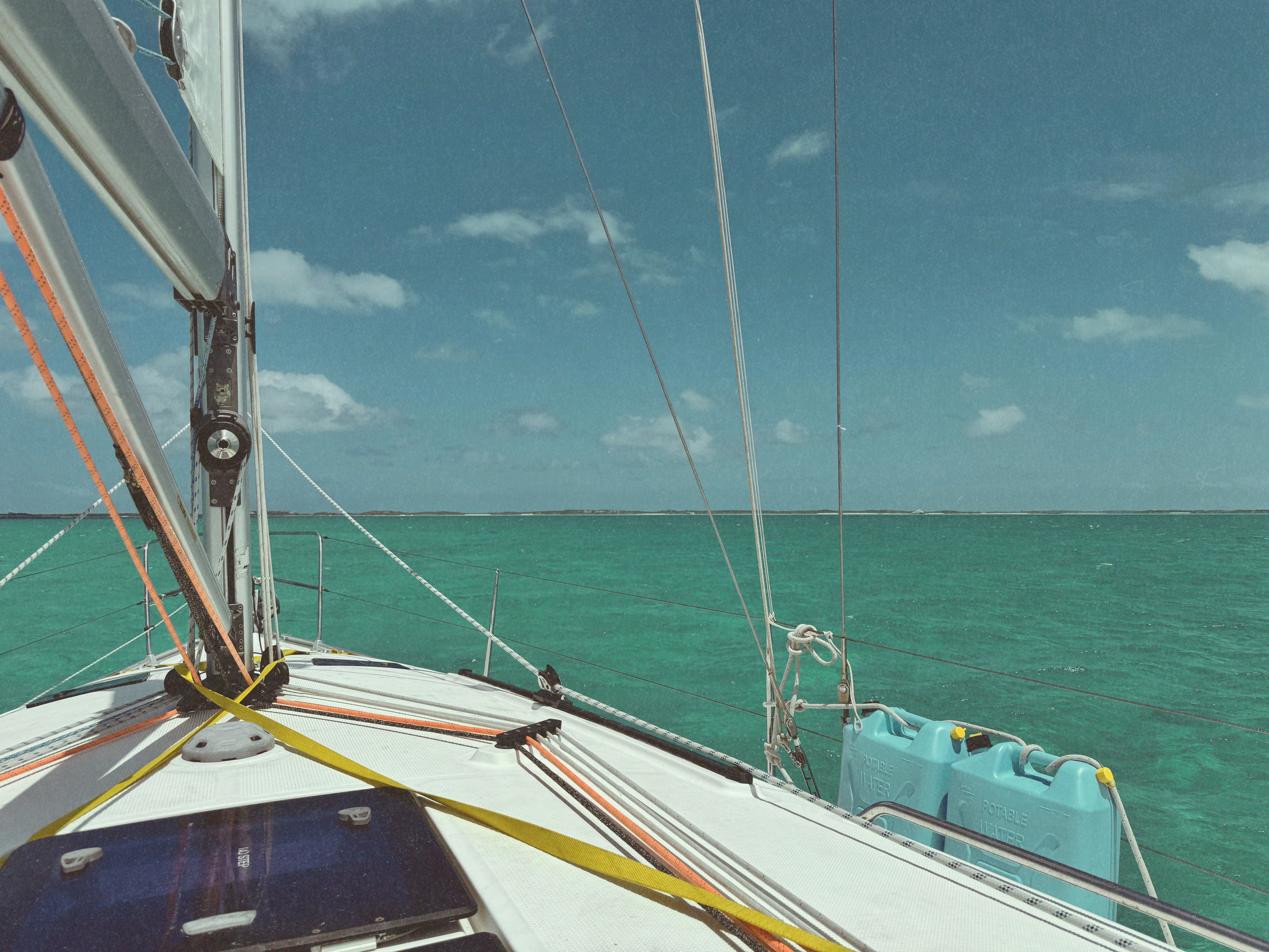 The bow of a cruising sailboat running over a turquoise Bahamian bank under a bright sky, with a low cay on the horizon.